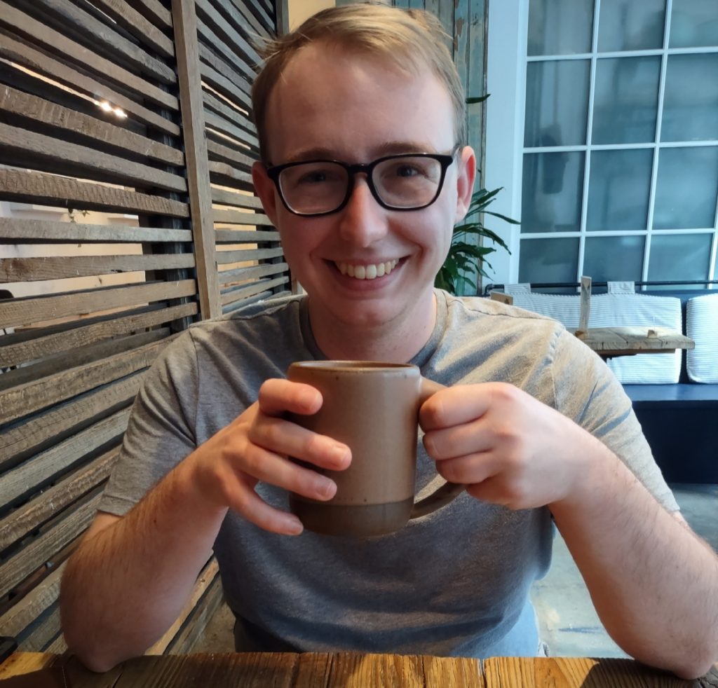 ITEHP student, Alex George, wearing a gray t-shirt and smiling over a cup of coffee