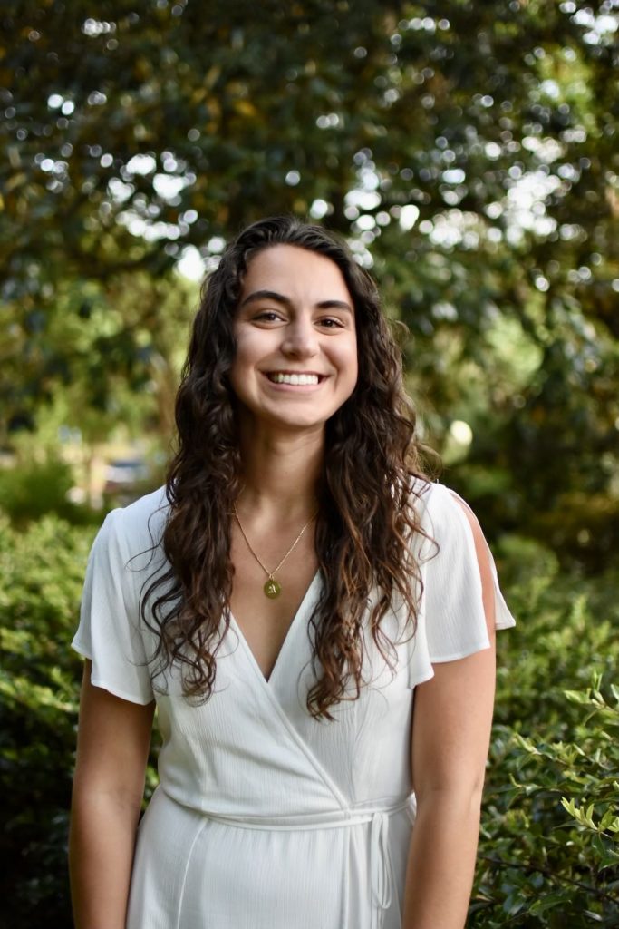 ITEHP student, Amelia Foley, smiling and wearing a white dress, standing in front of green, leafy trees