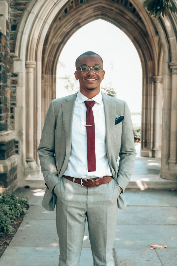 ITEHP student, Andrew Wrench, wearing a light gray suit with red tie, while standing outside under a Duke Chapel archway