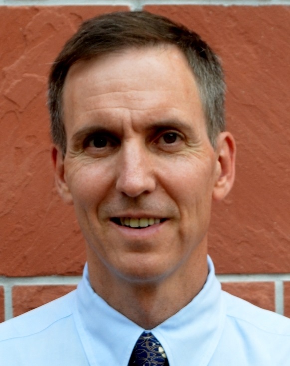 Jeff Burgess, MD, MS, MPH, smiling and wearing a collared shirt and tie while standing in front of a red brick wall