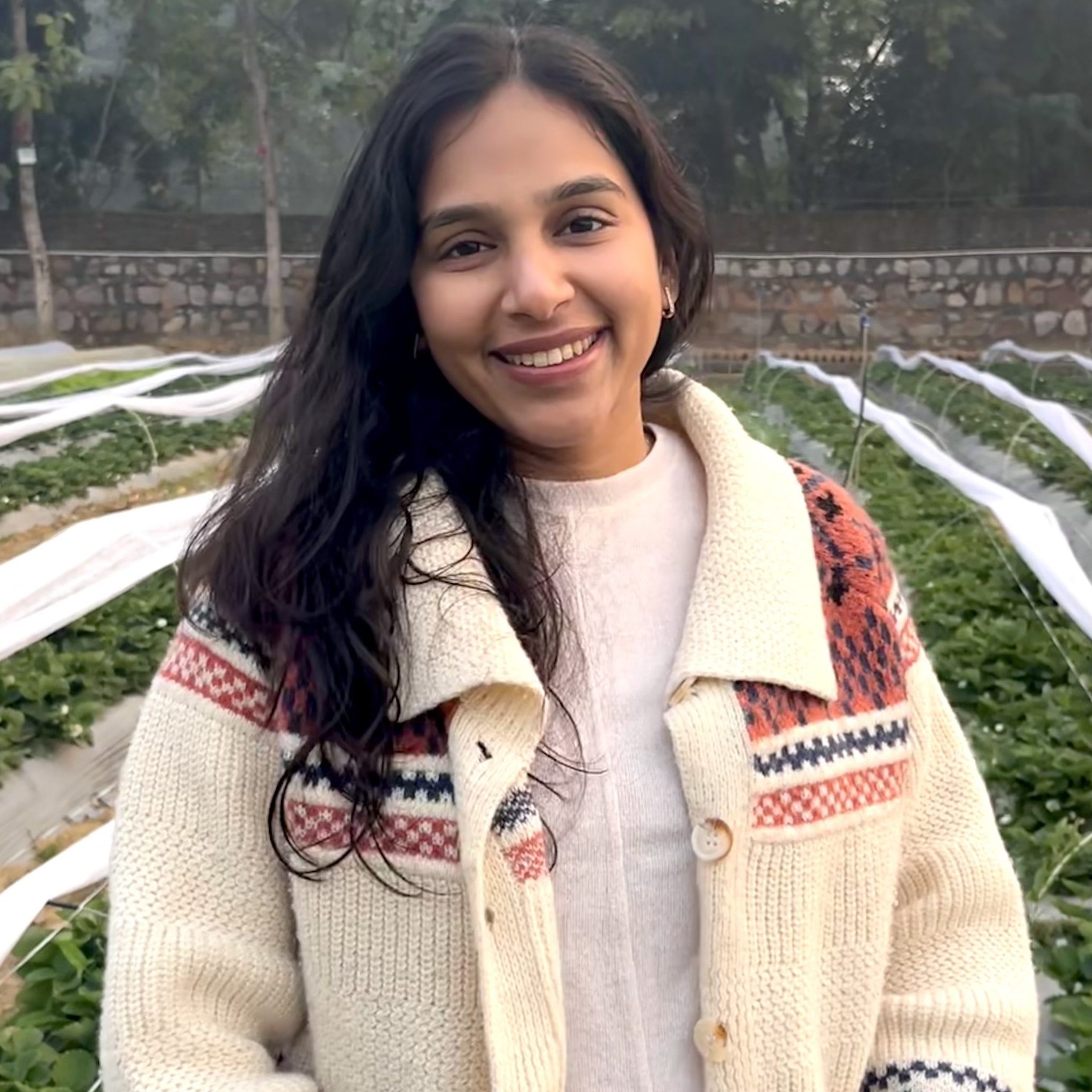 A person with brown skin and long, dark hair standing in a field of strawberry plants. She wears an off-white cardigan with a collar and some brown and orange patterns.