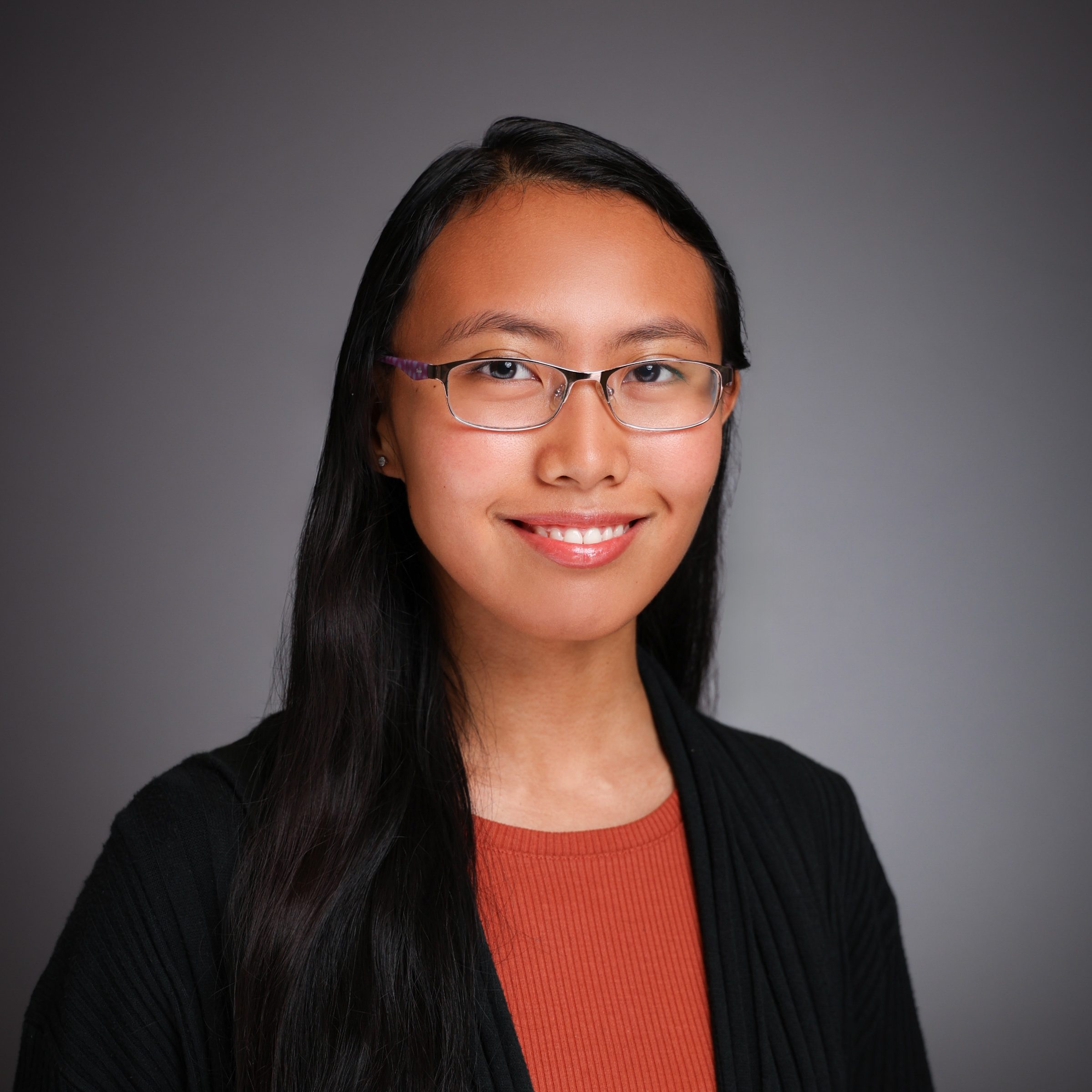 A studio portrait of a smiling woman with light brown skin, long, black hair, and metal-framed glasses wearing a red shirt with a black jacket.