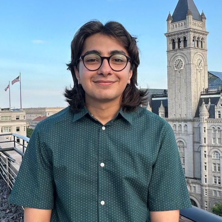A person with brown hair and medium tan skin smiling, wearing a dark green shirt and glasses, and standing outside with blue sky and buildings in the background